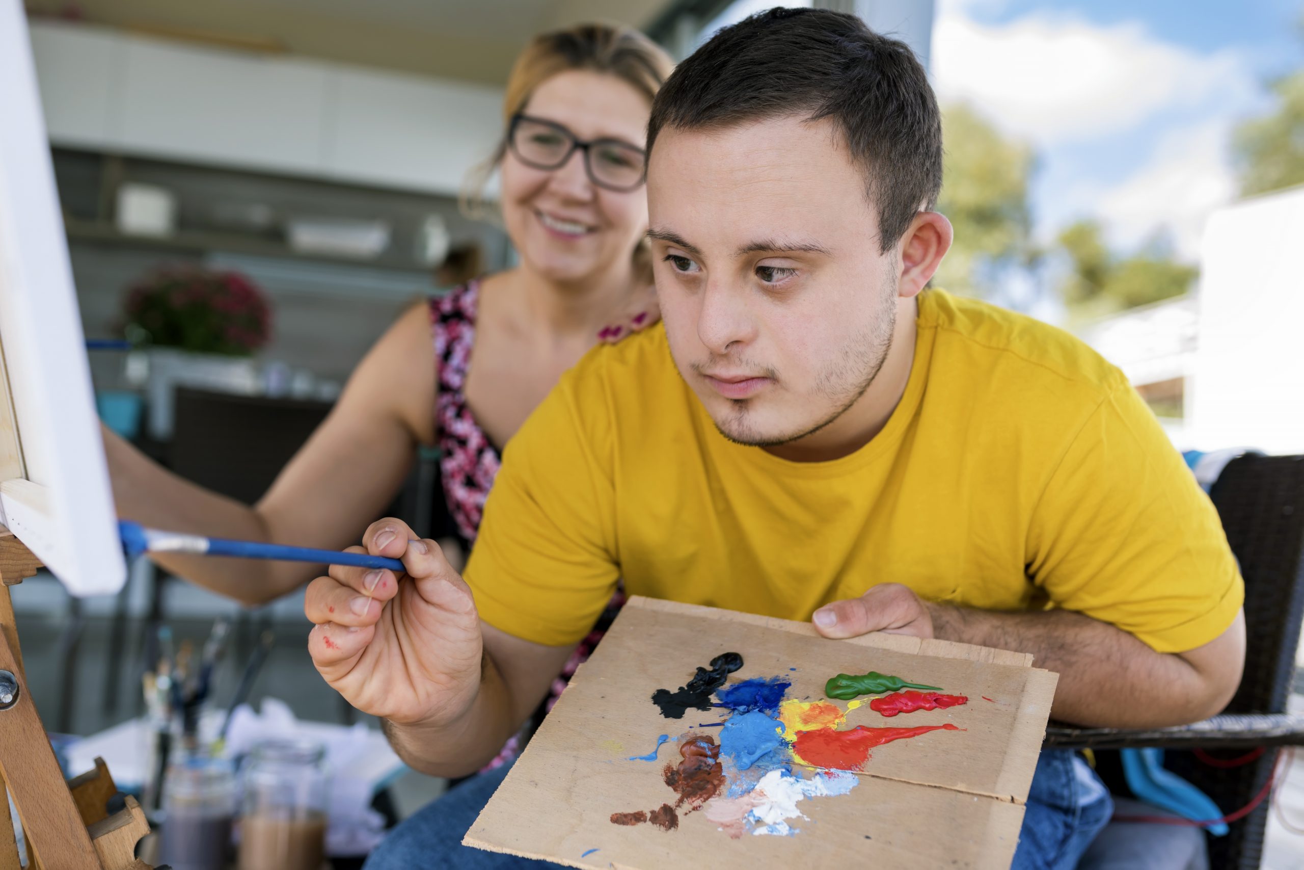 Teen boy with Down syndrome painting on canvas with his tutor    on terrace above riverbank